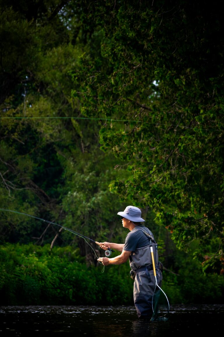 Man fly fishing in a lush green river landscape during daytime.